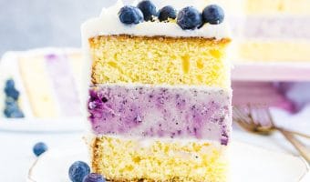 shot of a slice of lemon blueberry cheesecake layer cake sitting on a plate with the rest of the cake in the background