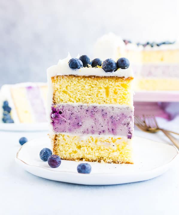 shot of a slice of lemon blueberry cheesecake layer cake sitting on a plate with the rest of the cake in the background