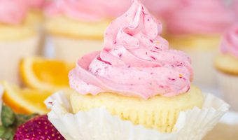 champagne cupcake with strawberry-orange buttercream (strawberry mimosa cupcakes) sitting on a plate with a sliced strawberry next to it and more cupcakes in the background