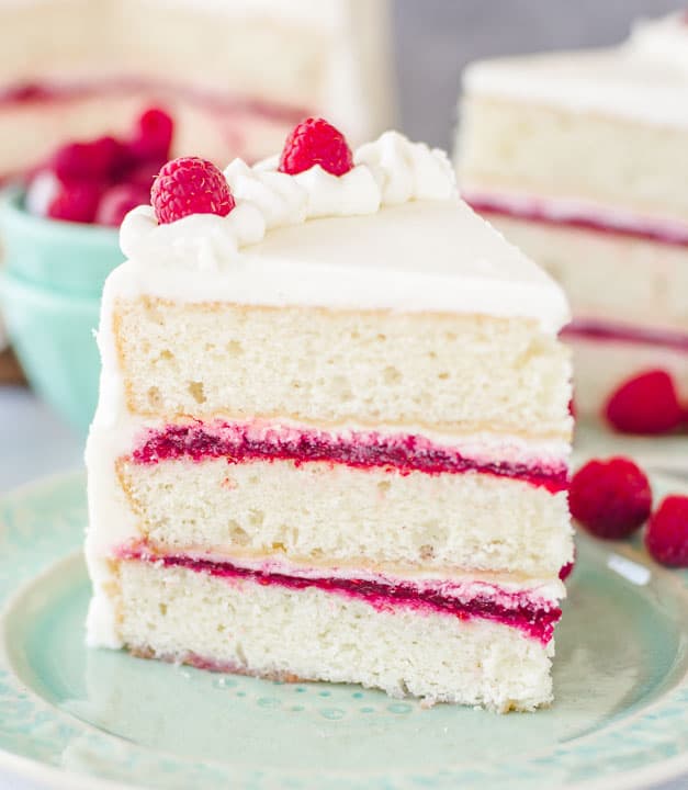 slice of white chocolate raspberry cake sitting on a plate with a bowl of raspberries behind it