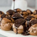 chocolate profiteroles sitting on a tray with a pitcher of glaze in the background