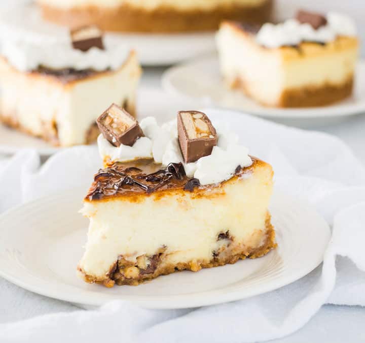slice of white chocolate snickers cheesecake on a plate with the rest of the cheesecake in the background