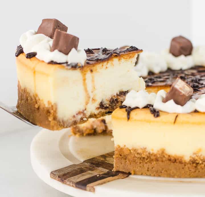 white chocolate Snickers cheesecake on a cake plate with a slice being lifted off of it