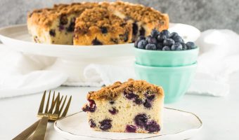 slice of blueberry buckle on a plate with the rest of the cake in the background on a stand