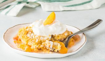 fresh peach dump cake on a plate with a fork taking a bite out of it and the rest of the cake in the background