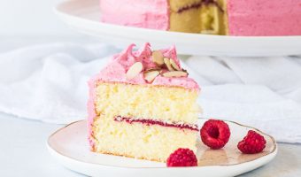 slice of raspberry almond cake on a plate with three raspberries and the rest of the cake in the background