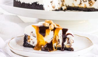 slice of Snickers ice cream cake on a plate with the rest of the ice cream cake behind it
