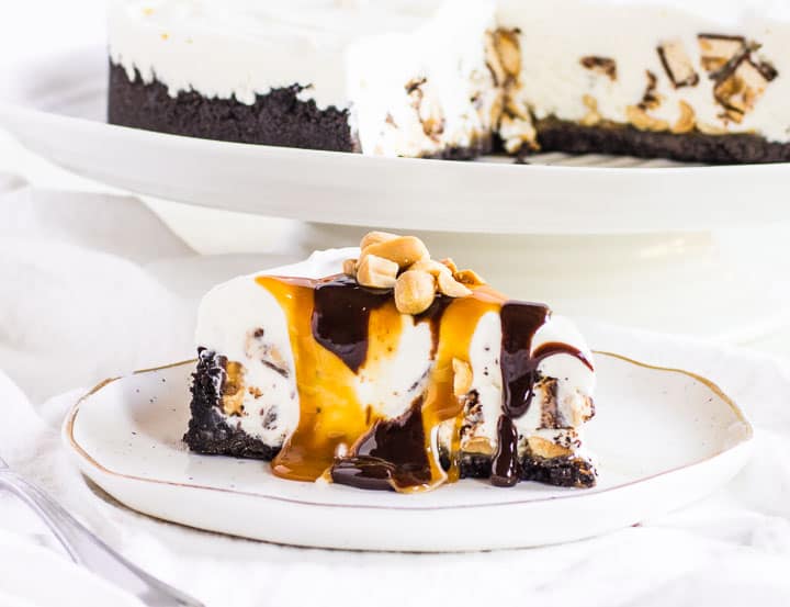 slice of Snickers ice cream cake on a plate with the rest of the ice cream cake behind it
