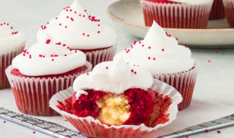 frosted red velvet cheesecake cupcakes on a wire rack with the front cupcake cut in half to show the filing
