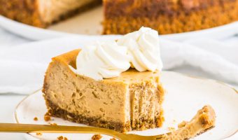 slice of cheesecake with a fork on a plate with the rest of the cheesecake on a cake plate in the background