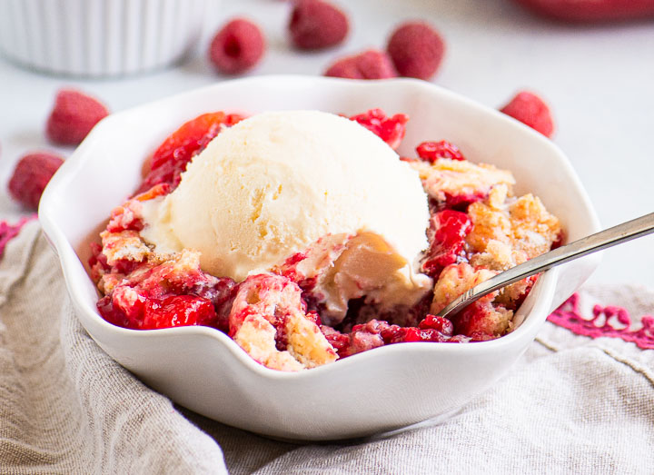 peach raspberry cobbler in a bowl with ice cream on top and raspberries scattered behind it