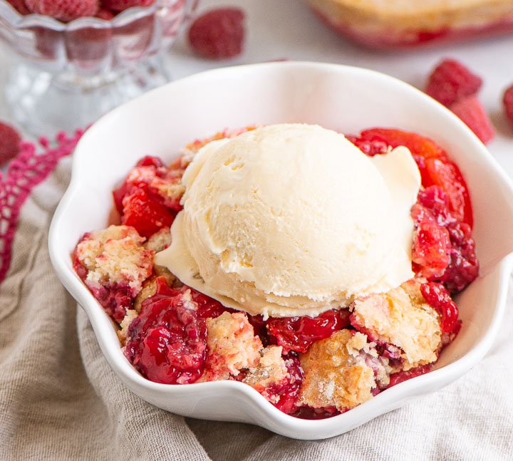 cobbler topped with vanilla ice cream in a bowl with a dish of raspberries behind it