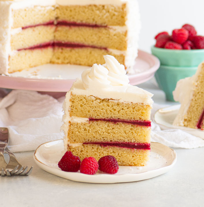 slice of cake on a plate with raspberries in front of the sliced cake on a cake plate