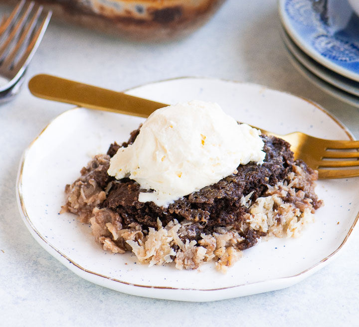 A serving of German chocolate dump cake topped with a scoop of vanilla ice cream on a white plate with a gold fork.