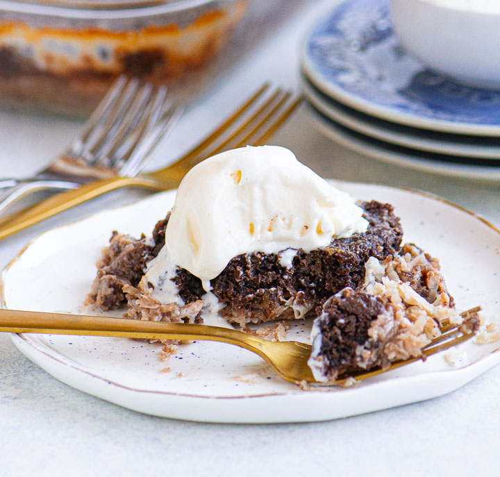 Close-up of a warm chocolate-coconut dump cake with vanilla ice cream melting on top, served on a white plate with gold flatware.