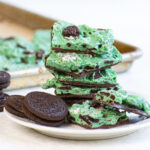 Several pieces of green peppermint Oreo bark stacked on a white plate, with whole Oreo cookies on the side and more bark cooling on a tray in the background.